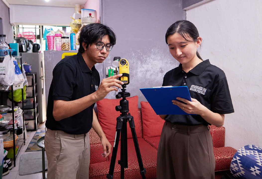 Two SUTD students using a yellow sensor on a tripod, with one taking notes on a clipboard.